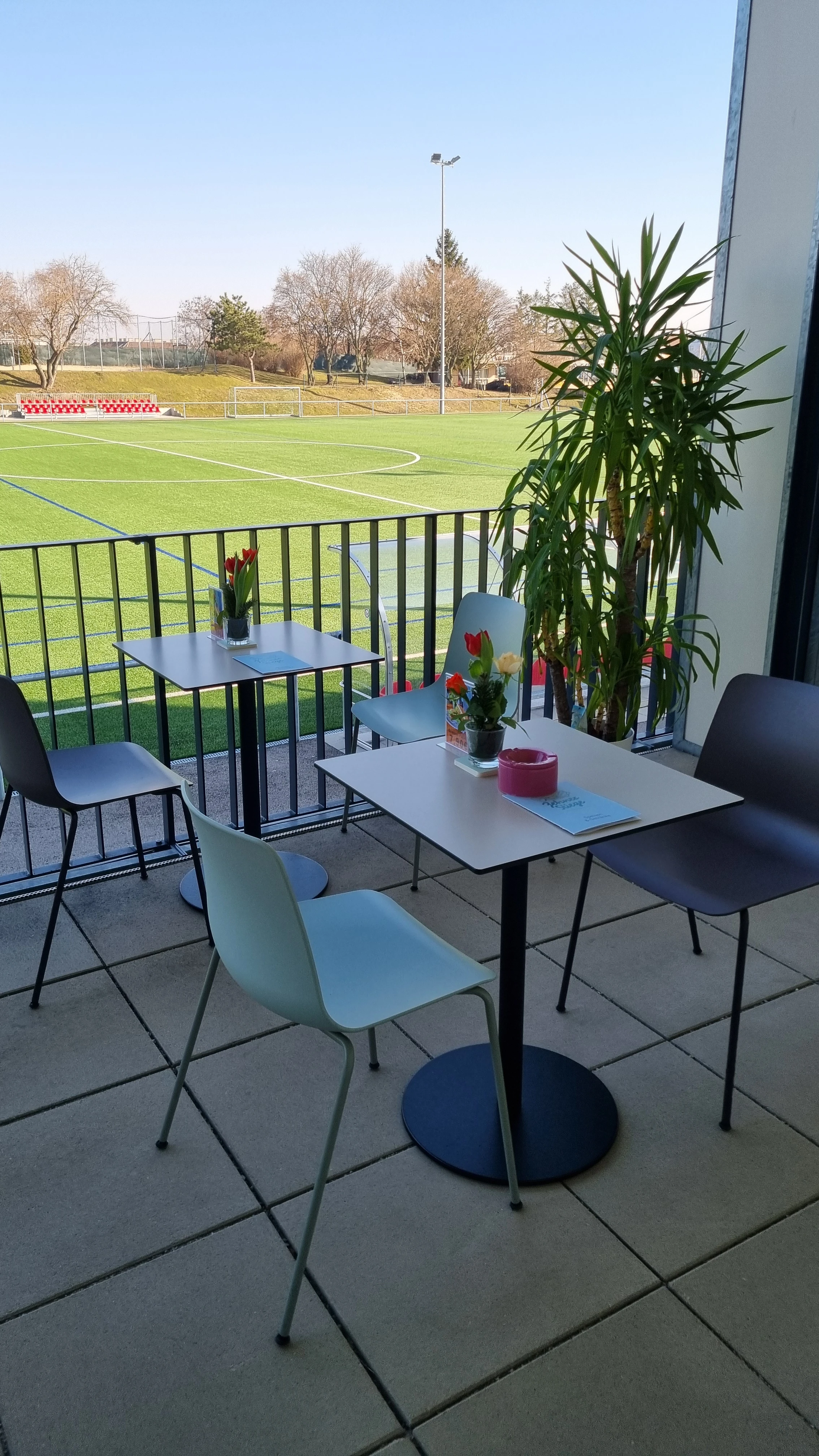 Sonnige Terrasse mit Blick aufs Fußballfeld; weiße Tische & grau/hellgraue Stühle.